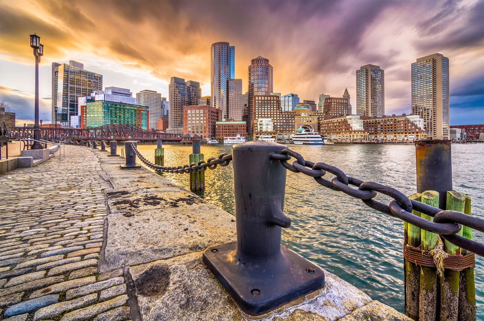 Cobblestone harbor walk with iron bollards and chains facing the Boston skyline at dusk